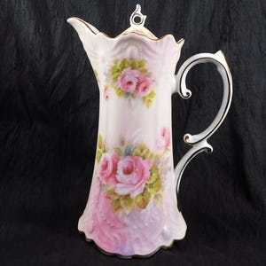 Può includere: Brocca alta e ornata in porcellana bianca e rosa, con rose rosa dipinte a mano e foglie verdi. La brocca ha un manico curvo e dettagli dorati, con un bordo smerlato e un pomello decorativo sul coperchio. Alta circa 25 cm.