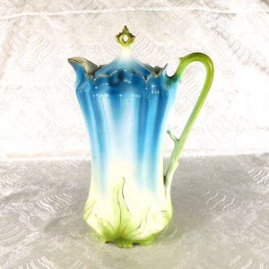 May include: A porcelain jug featuring a vertical fluted body with a blue-to-white gradient finish. The base is decorated with green leaf-like accents, and the handle is shaped like a green stem. It includes a matching lid with a finial.