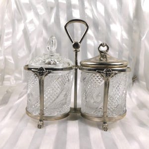 Op de afbeelding: Een vintage glazen en zilveren condimentenset. De set bevat twee heldere, glazen potten met zilveren deksels en decoratieve handvatten. De potten worden in een zilveren metalen standaard met een handvat gehouden. De achtergrond is een witte, getextureerde stof.