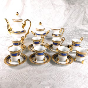 Puede incluir: Juego de té de porcelana blanca con ribetes dorados y detalles azules. El juego incluye una tetera, azucarero, jarrita para la leche y varias tazas con platillos. Las asas y detalles son dorados, creando un aspecto elegante.