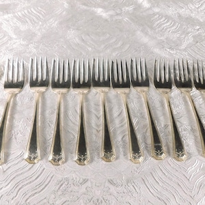 Puede incluir: Un juego de doce tenedores de metal plateado dispuestos en una fila horizontal. Cada tenedor presenta una cabeza de cuatro dientes y un mango delgado que se ensancha hacia la base, decorado con un sutil patrón floral en relieve en el extremo.