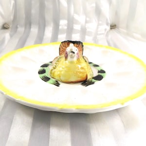 May include: A vintage ceramic egg plate with a yellow border and a central hen figurine. The hen is yellow, brown, and white, with black and green accents. The plate has indentations for holding eggs.