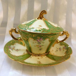 May include: A vintage porcelain tea cup, saucer, and lidded set. The set is decorated in green and cream with gold detailing. Floral patterns are painted on the cup, saucer, and lid, creating an elegant and refined appearance.