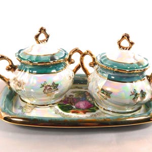 Op de afbeelding: Een vintage porseleinen suiker- en roomstel met een bijpassend dienblad. De set heeft iriserende witte, blauwgroene en gouden accenten. Elk stuk is versierd met bloemmotieven en sierlijke handvatten. De lade heeft een gouden rand.
