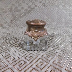 Puede incluir: Tintero de cristal antiguo con base cuadrada y tapa decorativa color bronce. La tapa y la base tienen detalles ornamentados. El tintero es transparente, lo que permite ver el contenido. Un accesorio de escritura clásico.