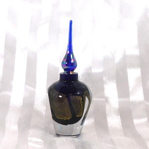 Puede incluir: Una botella de vidrio decorativa con un tapón azul largo y puntiagudo. La botella azul oscuro tiene detalles dorados y plateados. La botella tiene una forma y un diseño elegantes.