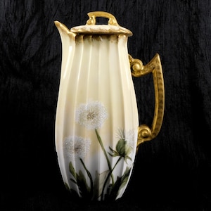 Puede incluir: Jarra de cerámica color crema con asa y tapa doradas. La jarra presenta un diseño acanalado y está decorada con flores de diente de león blancas pintadas a mano y hojas verdes. El asa y la tapa están adornadas con detalles dorados.