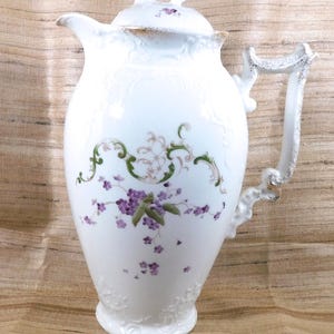 Puede incluir: Jarra de porcelana blanca con tapa, con un diseño floral en morado y verde. La jarra tiene un asa y un pico decorativos, con detalles dorados. El diseño incluye detalles en relieve y una estética delicada y ornamentada.