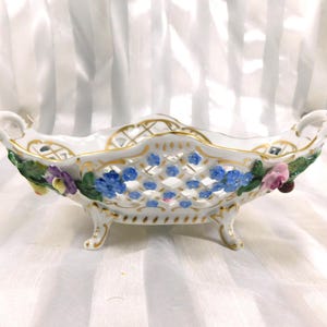 May include: A white porcelain serving dish with a lattice design and gold trim. The dish is decorated with blue and purple flowers and green leaves. It has two handles and sits on small feet.