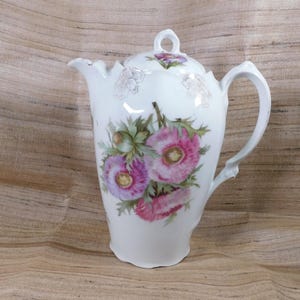 May include: White porcelain coffee pot with a lid, featuring a floral design of pink poppies and green leaves. The pot has a curved handle and a spout. The floral design is hand-painted on the pot's surface.