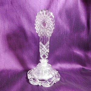 Puede incluir: Un frasco de perfume de cristal tallado transparente con un tapón decorativo. El frasco tiene una base redonda y festoneada y un tallo largo y ornamentado. El tapón tiene un diseño de sol. El fondo es de tela morada oscura.
