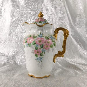 May include: A white porcelain pitcher with a gold handle and trim. The pitcher is decorated with pink and blue floral designs. The lid is topped with a gold finial. The pitcher has a scalloped rim.
