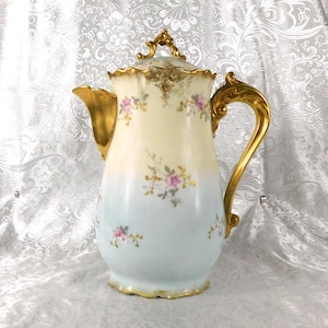 May include: An antique porcelain coffee pot with a gold handle, spout, and accents. The pot is painted with a gradient of colors, from cream to light blue, and adorned with pink floral designs. The lid is also gold and cream, with a decorative handle.