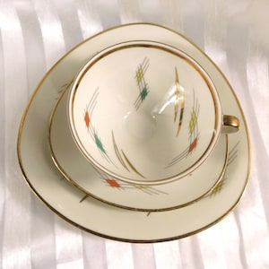 May include: A vintage cream-colored teacup, saucer, and plate set. The set features gold trim and a geometric design with red, green, and yellow accents. The teacup has a gold handle and sits on a matching saucer and plate.