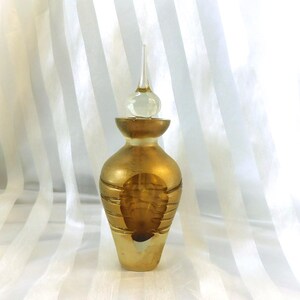 Könnte beinhalten: Dekorative goldfarbene Parfümflasche mit einem durchsichtigen Glasverschluss. Die Flasche hat eine bauchige Form mit horizontalen Goldstreifen und einem detaillierten Design. Der Verschluss ist transparent mit einer spitzen Spitze.