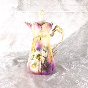 Może przedstawiać: Wysoki, zdobiony porcelanowy dzbanek z kwiatowym wzorem. Dzbanek posiada delikatny uchwyt i pokrywkę. Paleta kolorów obejmuje odcienie żółtego, różowego, fioletowego i białego. Dzbanek jest prawdopodobnie używany do serwowania napojów.