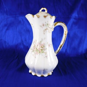 May include: A white porcelain pitcher with a fluted body and a matching lid. The pitcher features delicate pink floral patterns and gold trim along the handle, rim, spout, and lid. The base and top edges have a scalloped design.