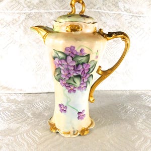 Può includere: Una caffettiera in porcellana dal corpo color crema, decorata con un mazzetto di violette viola e foglie verdi dipinte a mano. Il recipiente presenta un manico dorato, un coperchio abbinato con pomello dorato e bordi smerlati in oro.