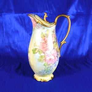 May include: A porcelain pitcher featuring a cream-colored base with hand-painted pink rose clusters and green foliage. The vessel includes a gold-toned handle, a scalloped gold rim, and a decorative gold base, showcasing a curved, elegant silhouette.