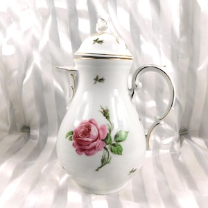 Puede incluir: Cafetera de porcelana blanca con un diseño de rosa rosa. La cafetera tiene tapa, asa curva y ribete dorado. También hay hojas verdes y pequeños detalles florales. Diseño clásico.