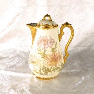 Op de afbeelding: Crèmekleurige porseleinen koffiepot met een gouden handvat, tuit en rand. De pot is versierd met roze, perzik en gele bloemmotieven. Het deksel is ook crèmekleurig met gouden rand en een klein gouden handvat.