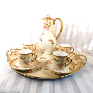 Puede incluir: Un juego de té vintage con una cafetera de porcelana color crema, cuatro tazas y platillos, todos adornados con delicados diseños florales rosas y detalles dorados. El juego se presenta en una bandeja ovalada con asas ornamentadas.