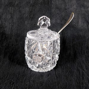 May include: A clear, cut-glass sugar bowl featuring a geometric diamond pattern across its cylindrical body and lid. The lid is topped with a faceted knob, and a silver-toned metal spoon rests inside the container, extending from the top opening.