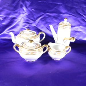 May include: A white porcelain tea set comprising a teapot, a lidded sugar bowl, a creamer, and a tall coffee pot. Each piece features a delicate floral pattern in pastel colours, accented with gold trim on the rims, handles, and lids.