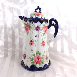 Op de afbeelding: Witte porseleinen kan met een deksel, versierd met roze en paarse rozen, groene bladeren en gouden accenten. De kan heeft een marineblauwe handgreep en rand met een geschulpte rand. Een bloemmotief bedekt het deksel.