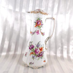 May include: White porcelain jug with a floral pattern in purple, pink, orange, and green. The jug has a tall, slender shape, a decorative handle, and a lid with gold trim. A classic piece for serving beverages.