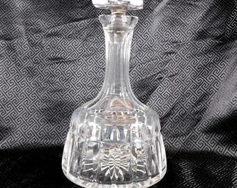 May include: A clear crystal glass decanter with a stopper. The decanter has a wide, faceted base that tapers to a slender neck and a decorative stopper. The glass reflects light, creating a sparkling effect. The decanter is likely used for serving wine or spirits.