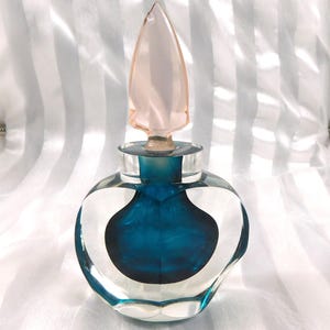 Puede incluir: Un frasco de perfume de vidrio transparente con un interior de color turquesa y un tapón puntiagudo rosa. El frasco tiene una forma redondeada con un diseño en forma de corazón en los lados. El tapón es de color rosa claro.