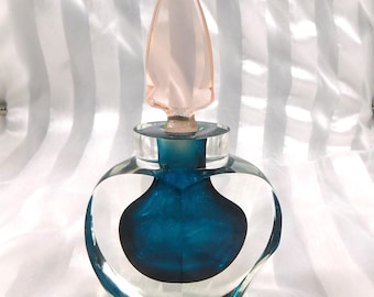 Könnte beinhalten: Eine transparente Parfümflasche aus Glas mit teal-farbenem Innenraum und einem rosa, spitz zulaufenden Verschluss. Die Flasche hat eine abgerundete Form mit einem herzförmigen Design an den Seiten. Der Verschluss ist hellrosa.