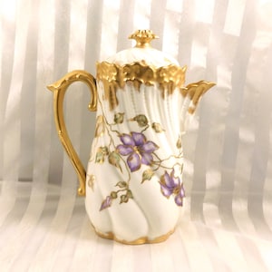 Può includere: Caffettiera in porcellana bianca con finiture e manico dorati. La caffettiera presenta un motivo floreale con fiori viola e foglie verdi. Il coperchio è sormontato da un pomello dorato. La caffettiera ha un design classico ed elegante.