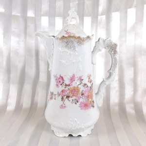 以下が含まれることがあります： ピンク、オレンジ、茶色の花柄模様の白い磁器コーヒーポット。装飾的なハンドルと蓋があり、金と銀のアクセントが付いています。デザインには、エンボス加工された花柄とスクロールワークのディテールが含まれています。