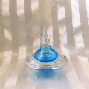 Può includere: Un flacone di profumo in vetro trasparente con interno blu e un lungo tappo a punta. Il flacone ha una base arrotondata e una parte superiore bulbosa, con un tappo in vetro trasparente. Il design è elegante e decorativo.