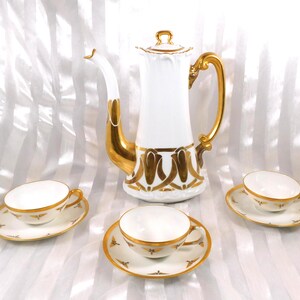Puede incluir: Una cafetera de porcelana blanca con detalles dorados, acompañada de tazas y platillos a juego. La cafetera tiene un asa y un pico dorados, con detalles dorados. Las tazas y los platillos tienen bordes dorados y patrones dorados decorativos.