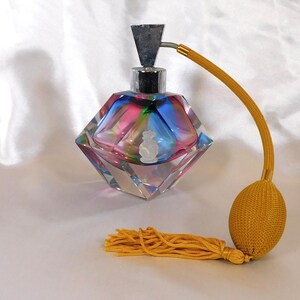 May include: A vintage glass perfume bottle with a geometric, faceted design. The bottle is filled with colorful liquid and has a silver atomizer. The bottle is attached to a long, yellow cord with a tassel and a yellow bulb at the end.