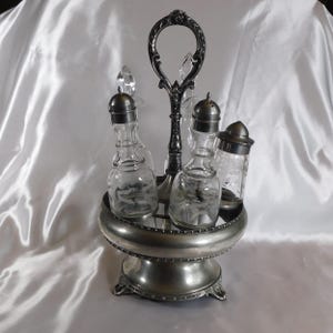 May include: A silver-toned condiment set with a handle, featuring two glass bottles with metal lids, a salt shaker, and a pepper shaker. The set is on a decorative, tiered base.