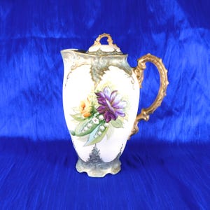 Può includere: Una brocca in porcellana con coperchio, caratterizzata da un motivo floreale dipinto a mano con fiori viola e gialli e foglie verdi. La brocca presenta un manico dorato ornato, una base smerlata e decorazioni dorate lungo il bordo.
