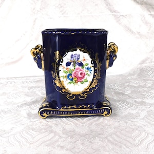 May include: A deep blue ceramic vessel featuring a central oval medallion with a colorful floral bouquet of pink roses and purple irises. The piece is accented with gold trim and two gold-colored ram head handles on the sides, set on a base.