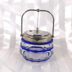 May include: A clear glass butter dish with a silver lid and handle. The glass has a blue stripe design around the middle and bottom. The handle is arched and attached to the lid and the base of the dish.
