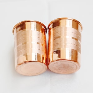 May include: Two copper drinking glasses with a hammered finish. The glasses have a shiny, metallic finish and are ideal for serving drinks.