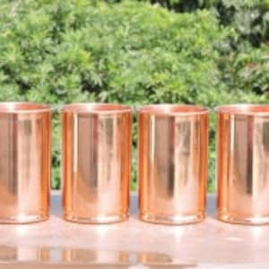 May include: Set of four copper drinking glasses, each with a smooth, shiny finish. The glasses are arranged in a row on a wooden surface.