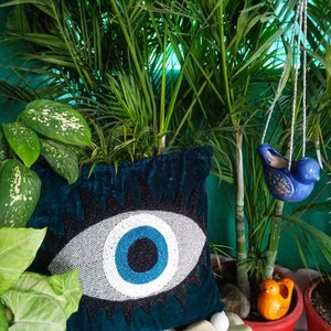 May include: Teal velvet pillow with a beaded evil eye design. The eye is white with a blue center and black outline. The pillow is surrounded by green plants and white stones.
