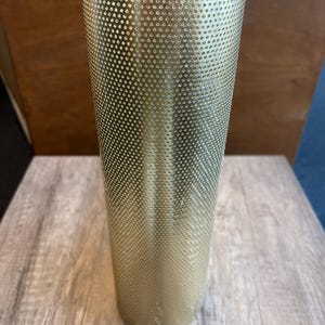 Może przedstawiać: Cylindryczny, złoty metalowy klosz z perforowanym wzorem. Klosz jest wysoki i smukły, z lekko rozszerzonym górnym brzegiem. Powierzchnia pokryta jest równomiernie rozmieszczonymi małymi otworami, tworzącymi teksturowany wygląd. Klosz jest prawdopodobnie do lampy stołowej lub podłogowej.