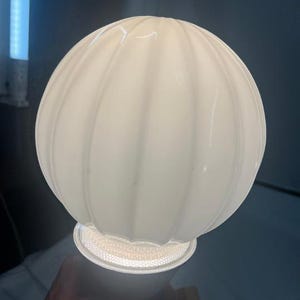 May include: A spherical, white, ribbed glass lamp shade with a translucent finish. The shade features vertical ridges that create a textured, rounded appearance. It is illuminated from within, casting a soft, even glow across the surface.
