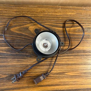Könnte beinhalten: Ein schwarzer und silberner Scheinwerfer mit einer Glühbirne, mit Netzkabel und Stecker. Der Scheinwerfer befindet sich vor einem hölzernen Hintergrund.