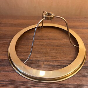 Puede incluir: Un anillo de pantalla de lámpara de color latón con un diseño circular. El anillo tiene un soporte de metal curvado que se conecta a una pequeña pieza superior circular. El anillo de la pantalla de la lámpara está sobre una superficie de madera.