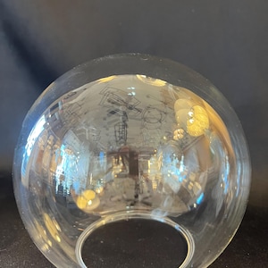 Op de afbeelding: Een helder, bolvormig glazen object met een cirkelvormige opening. Het glas reflecteert de omgeving en toont een vervormd beeld van de kamer. Het object heeft een gladde, afgeronde vorm.